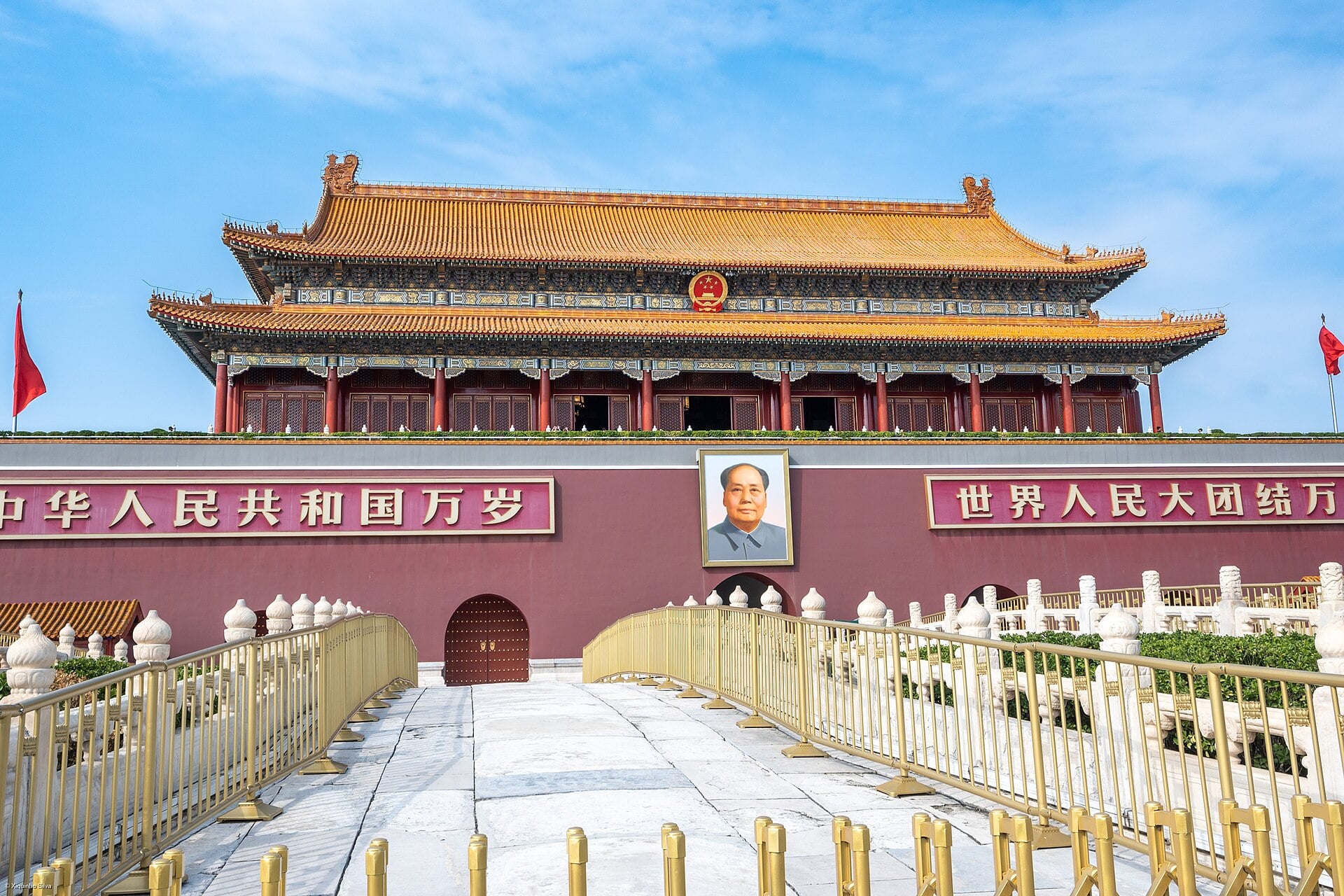 Tiananmen Gate in Beijing under a blue sky, with golden roof, red walls, portrait of Mao and Chinese banners, viewed from a fenced stone walkway