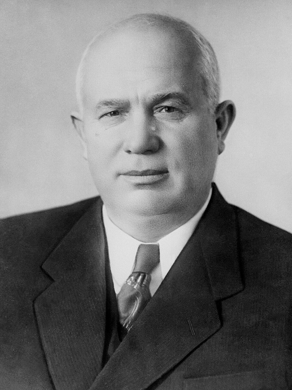 Black-and-white studio portrait of a middle-aged man in a dark suit and tie, facing the camera with a neutral expression against a plain background