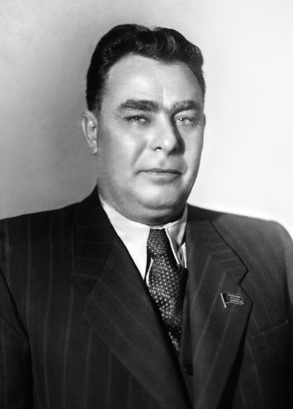 Black-and-white portrait of a middle-aged man in a pinstripe suit and tie, wearing a small lapel badge, facing the camera against a plain background