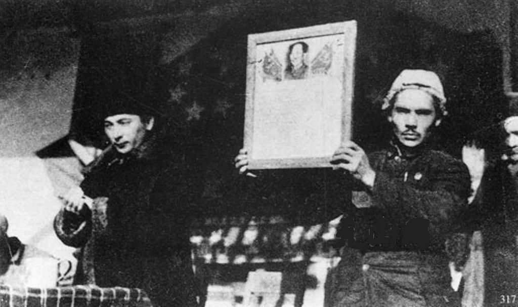 Historic black-and-white scene of a man holding a framed portrait while another man beside him gestures as if addressing an audience indoors