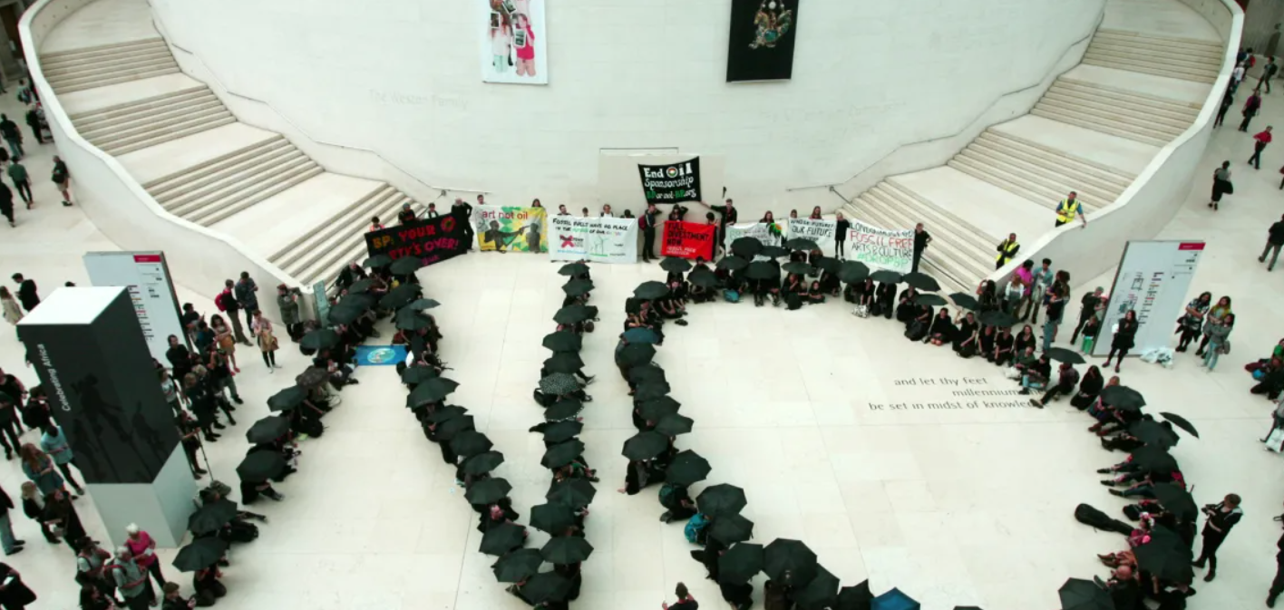 Protest in a large museum atrium where people with black umbrellas form the letters “NG”, holding banners opposing oil company sponsorship of the arts