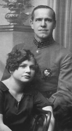 Vintage black-and-white portrait of a uniformed man seated behind a young woman, both facing the camera in a formal studio setting.