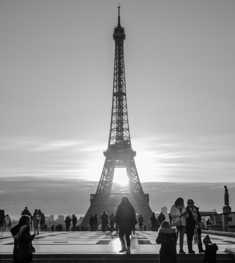 Black and white view of the Eiffel Tower at sunrise, silhouetted against the sky, with people walking and gathering on a plaza in the foreground