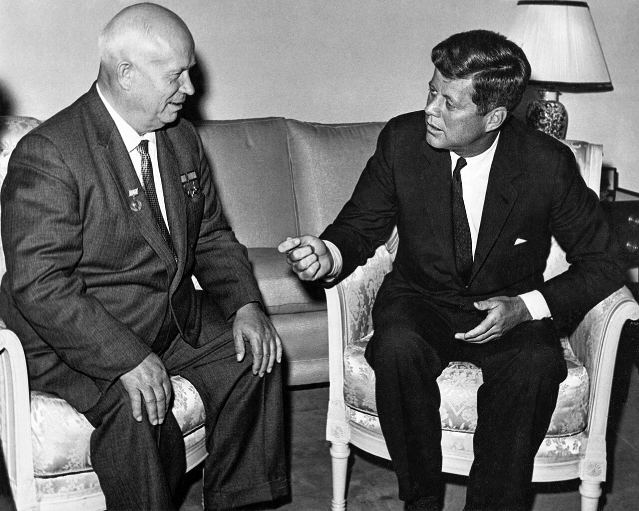 Black-and-white photo of two suited men smiling as they shake hands outside a formal building, surrounded by other officials watching closely