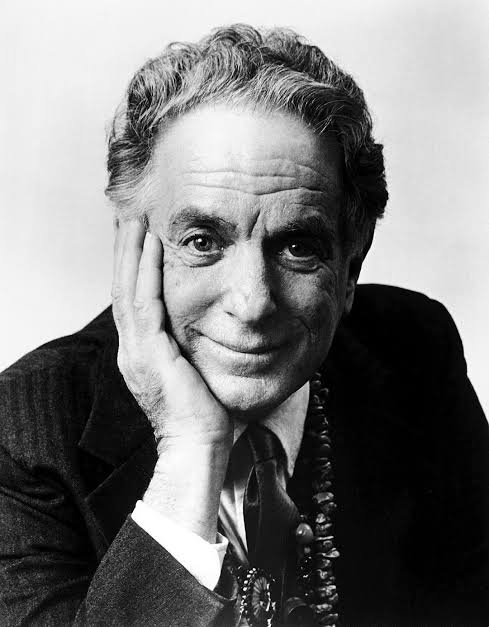Black-and-white portrait of an older man in a suit, smiling gently with his chin resting on his hand against a plain background