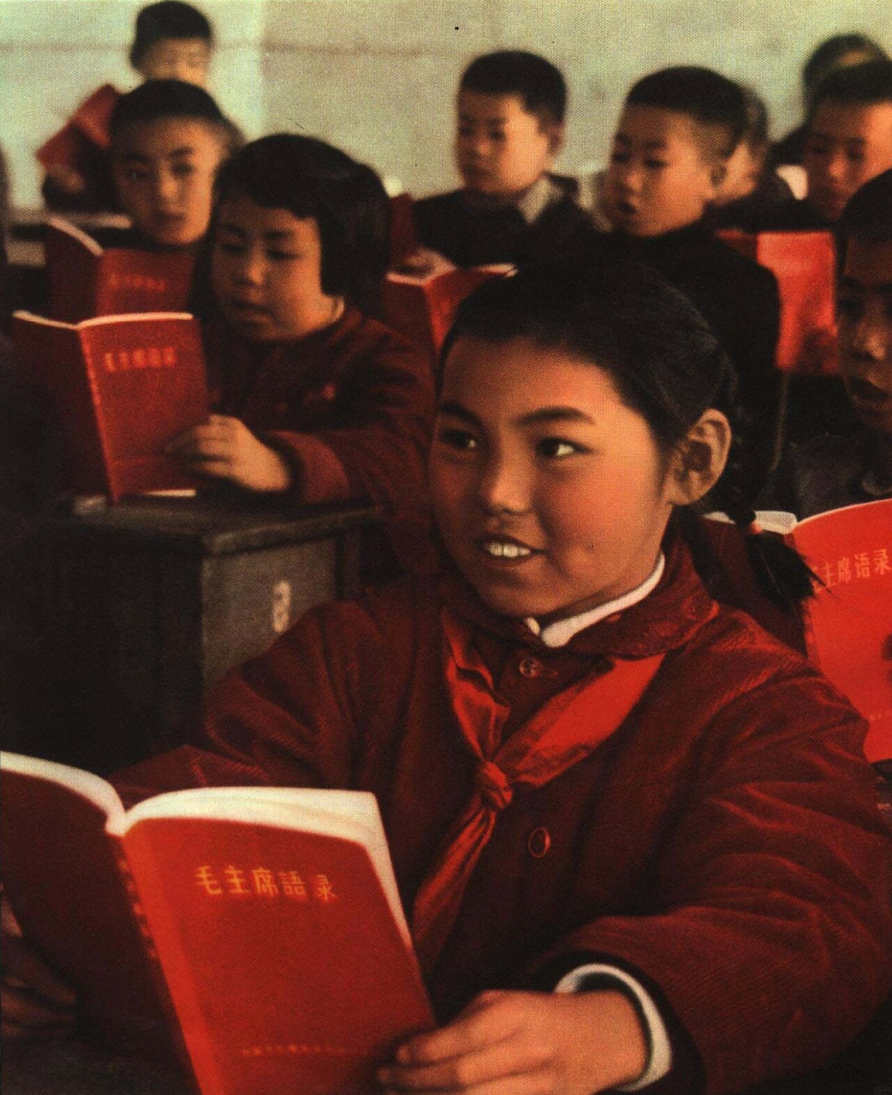 Chinese schoolchildren in uniform sit at wooden desks, reading from identical small red books in a classroom, with a girl in front smiling attentively