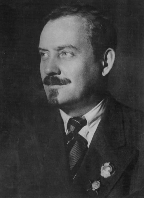 Black-and-white portrait of a middle-aged man in a suit and tie, with short hair, moustache and goatee, wearing two lapel badges and gazing left