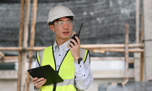 Construction site engineer in high-visibility vest and hard hat uses a walkie-talkie while holding a tablet, with scaffolding visible in the background.
