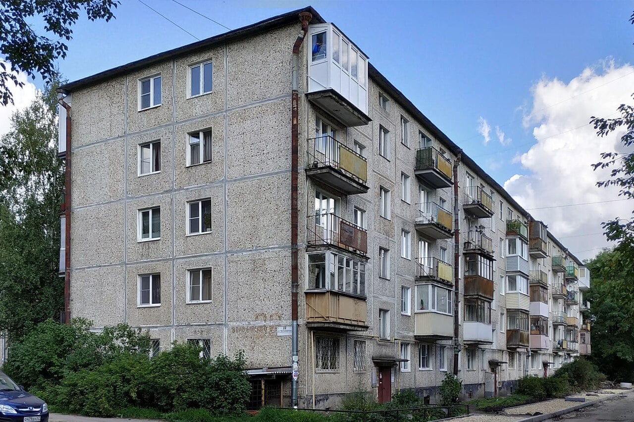 Old four-storey concrete block of flats with small balconies and overgrown shrubs at the base, lining a quiet street under a bright partly cloudy sky