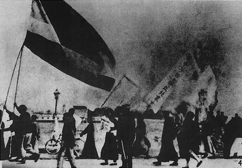Historic black-and-white photo of protesters marching with large flags in a smoky street, figures in dark clothing silhouetted against a hazy background