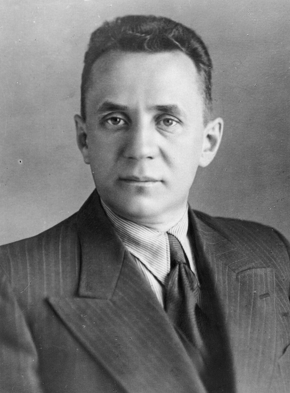 Black-and-white studio portrait of a middle-aged man in a pinstriped suit, shirt and tie, looking straight at the camera against a plain backdrop.