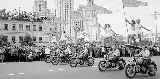 Women in leotards balanced on moving motorbikes wave flags in a city parade, while large crowds watch against a backdrop of tall Soviet-era buildings