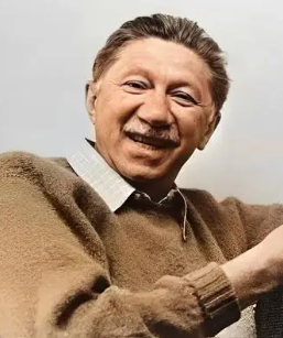 Smiling middle-aged man with short dark hair and moustache, wearing a brown jumper over a collared shirt, sitting relaxed against a plain light background