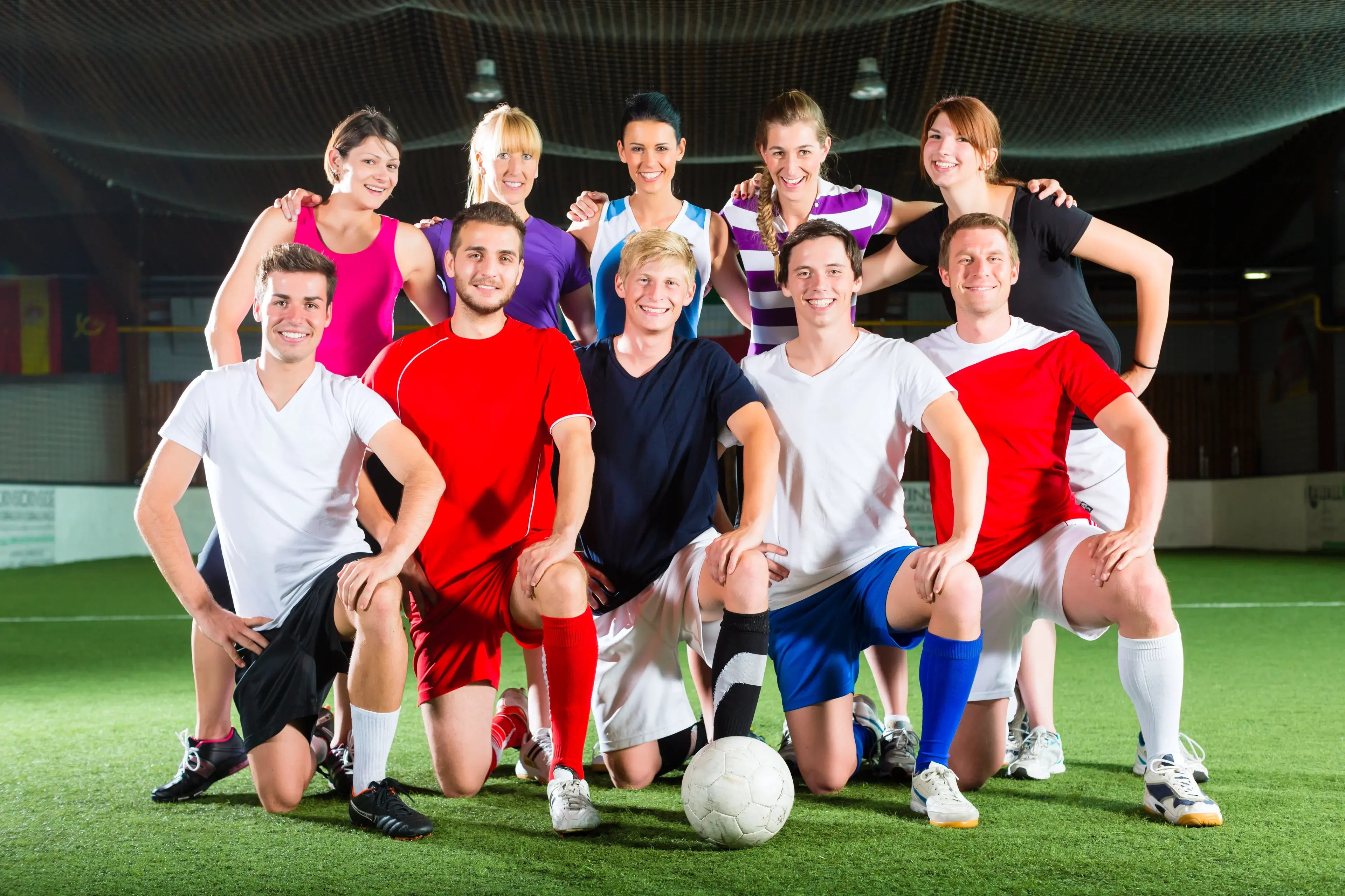 Mixed-gender five-a-side football team posing on an indoor pitch, with four kneeling players around a ball and six teammates smiling behind them