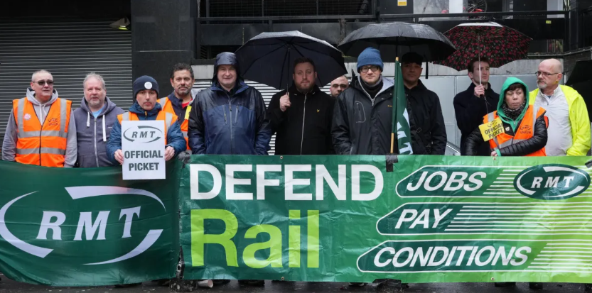 Rail workers stand in the rain behind an RMT banner reading “Defend Rail, Jobs, Pay, Conditions” during an official picket demonstration