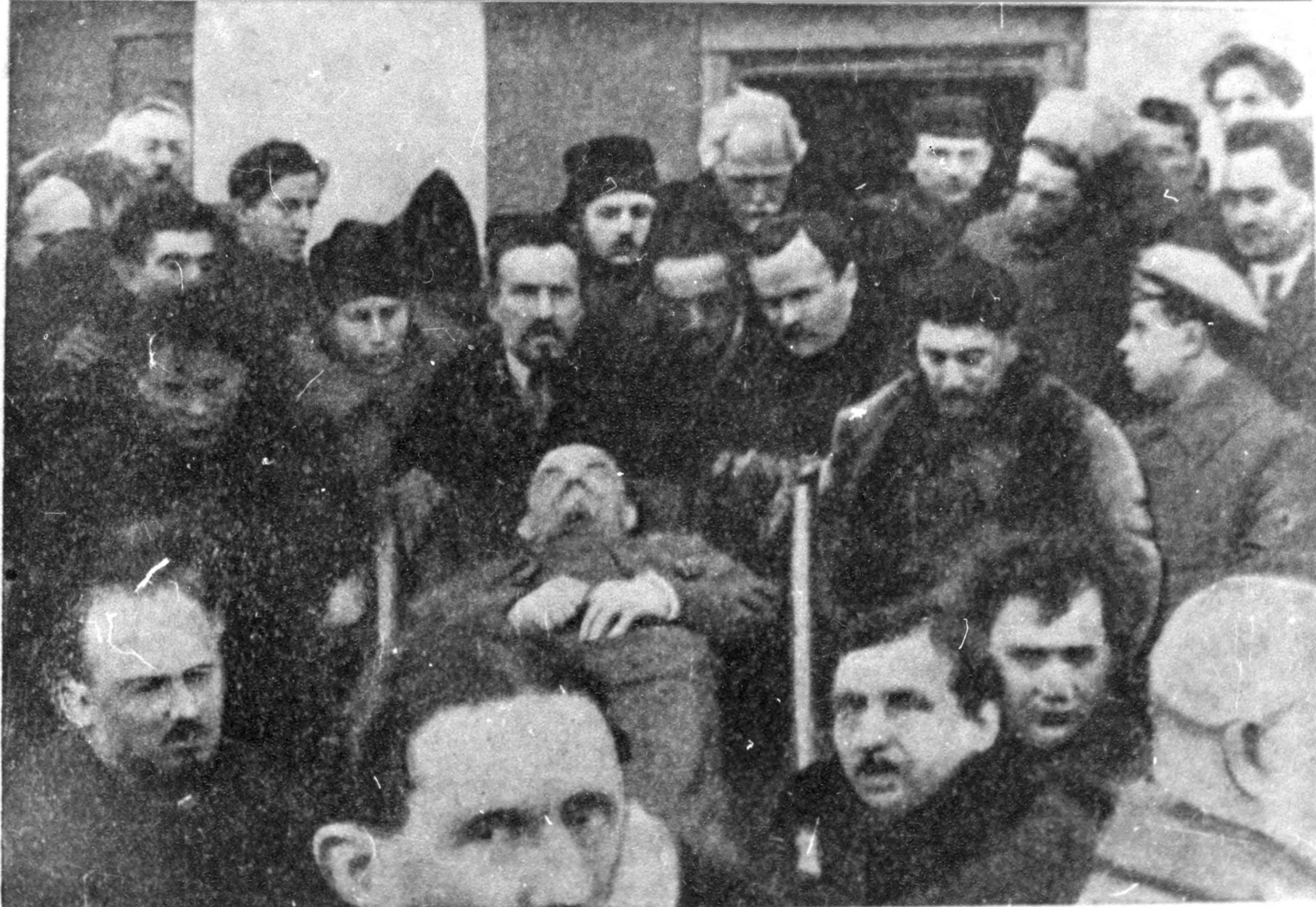 Historic black-and-white scene of solemn men gathered closely around a stretcher bearing an unconscious or deceased man in cold-weather clothing.