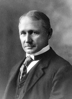 Black-and-white portrait of a middle-aged man in a suit, waistcoat and tie, with short parted hair and a neat moustache, looking calmly at the camera