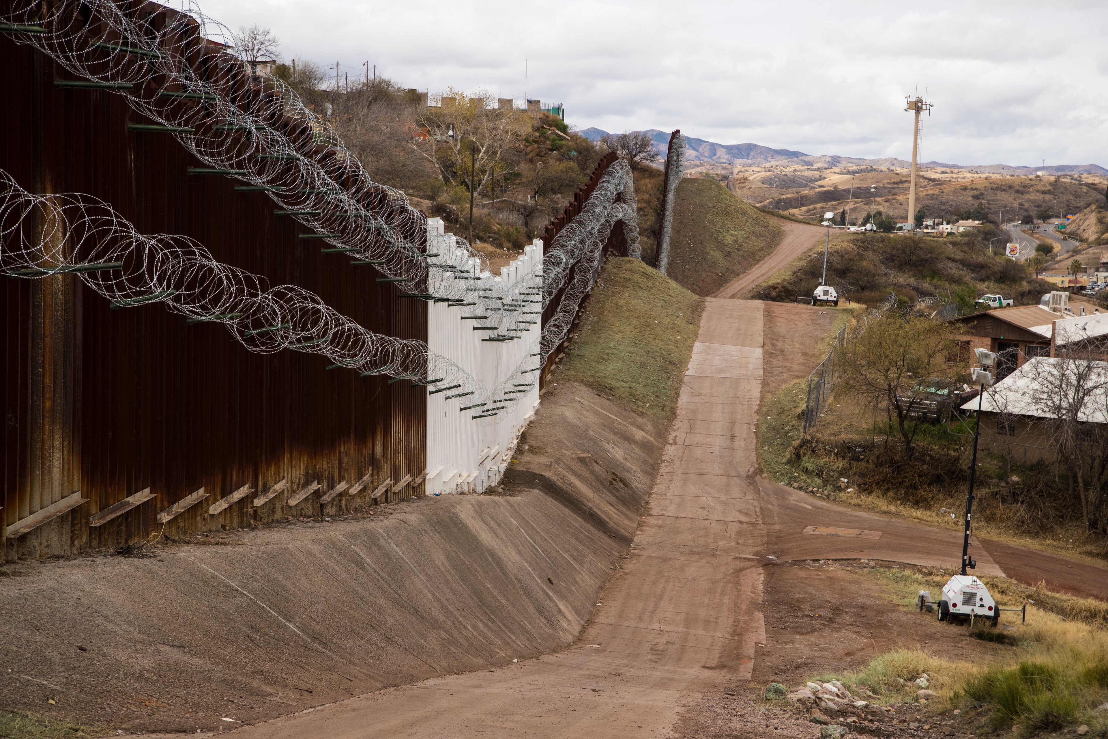Border wall lined with razor wire beside a dirt patrol road, with hills, scattered houses and security towers under a cloudy sky in the distance.