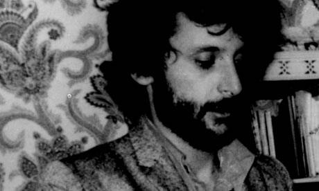 Black-and-white photo of a bearded man in a jacket, looking down thoughtfully in front of patterned wallpaper and a small bookshelf.