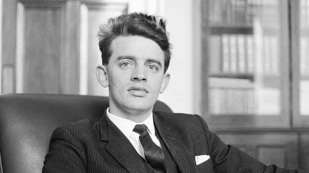 Black-and-white portrait of a young man in a suit and tie sitting in an office chair, with bookshelves and wooden panelling in the background