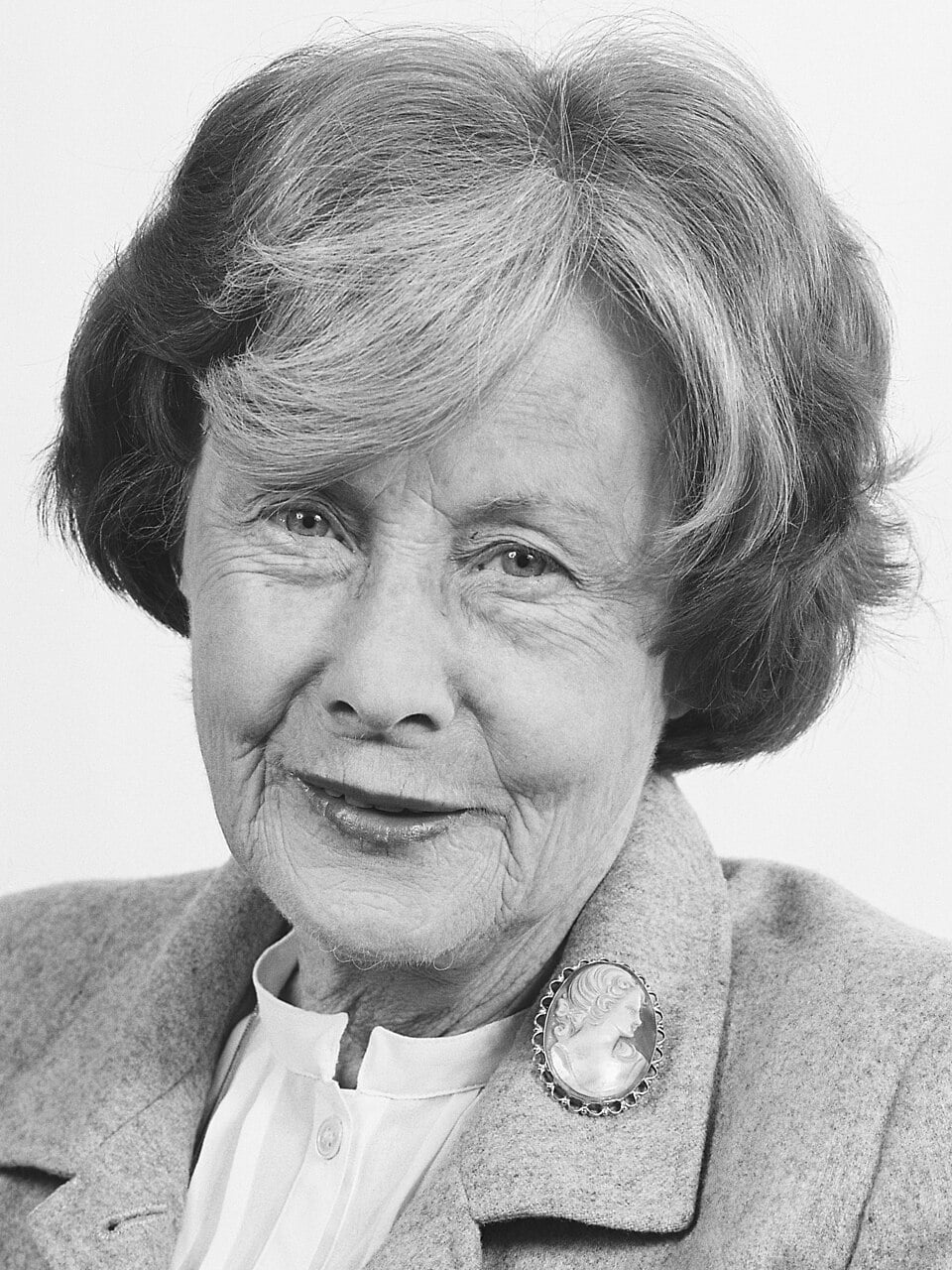 Black-and-white portrait of an elderly woman smiling gently, wearing a light coat, blouse, and large cameo brooch against a plain background