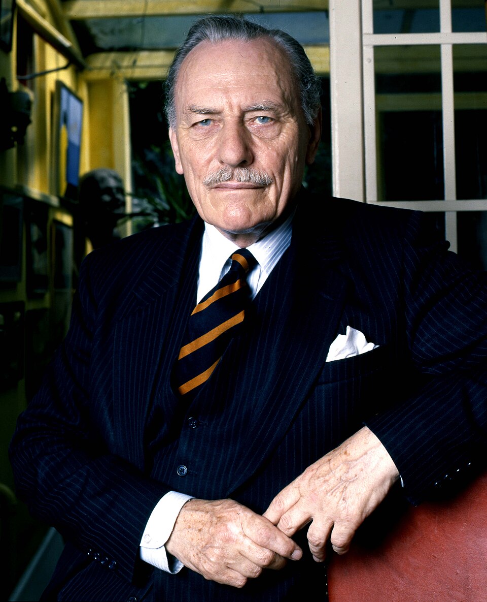 Elderly man in a dark pinstripe suit and striped tie stands indoors, hands folded, looking directly at the camera with a serious, composed expression