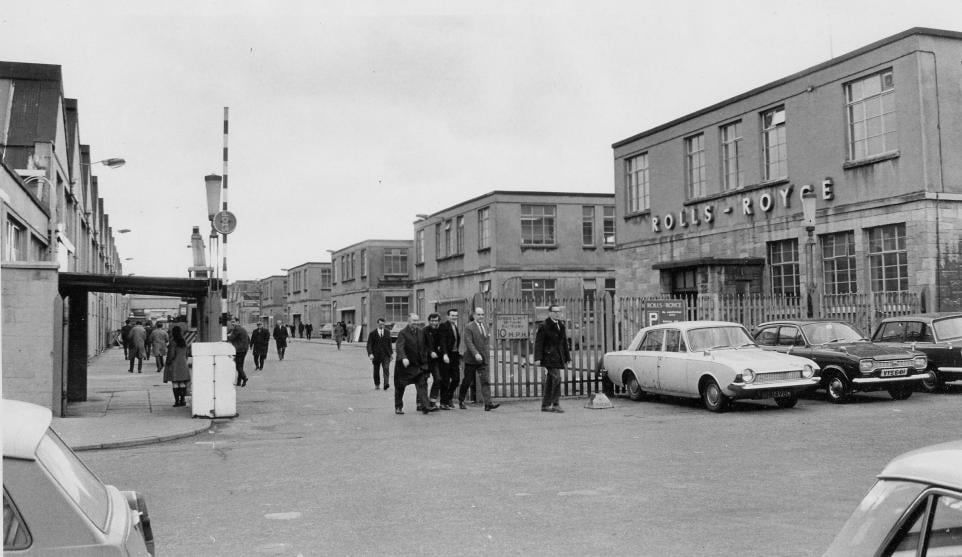 Black-and-white photograph of the Rolls-Royce factory, showing workers leaving through the gates, industrial buildings marked “ROLLS-ROYCE”, parked cars and a factory entrance sign.