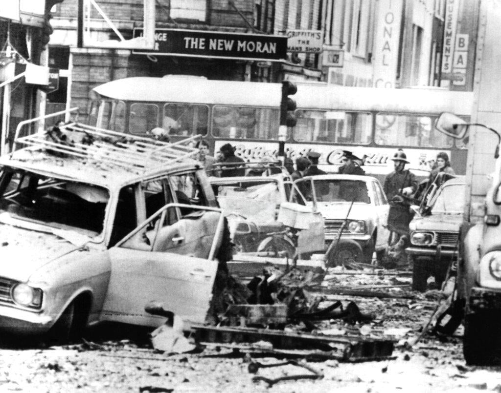 Black‑and‑white street scene showing bomb‑damaged cars, scattered debris and firefighters among wreckage outside shops and buses in a city centre