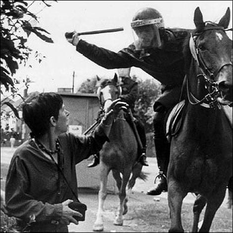Mounted riot police officer raising baton towards unarmed protester reaching up, in tense confrontation on a street, with another mounted officer behind.