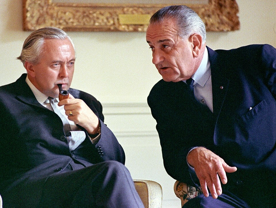 Two suited older men sit indoors in serious conversation, one leaning forward speaking as the other listens thoughtfully while holding a pipe.