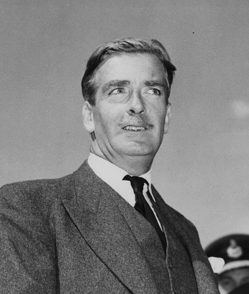 Anthony Eden in a suit and tie, with neat hair and moustache, looking slightly upward against a clear sky backdrop