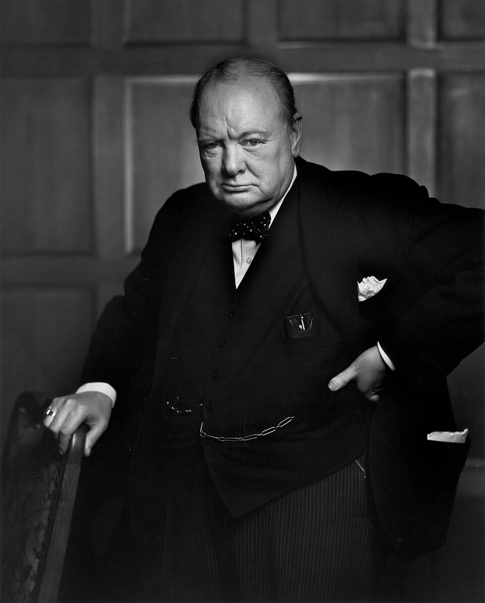 Elderly Winston Churchill in a dark suit and bow tie, standing in a wood-panelled room