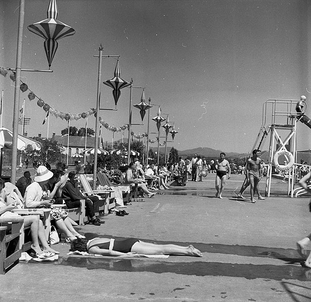 Crowded 1950s outdoor lido with people sunbathing, walking in swimwear and sitting on benches under decorative lamp posts, with hills visible in the distance