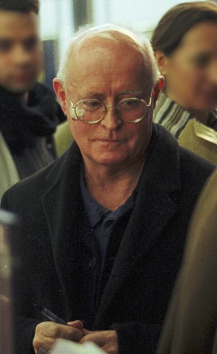 Older man with thinning grey hair and round glasses, wearing a dark coat, holding a pen in a busy indoor setting with blurred people in the background