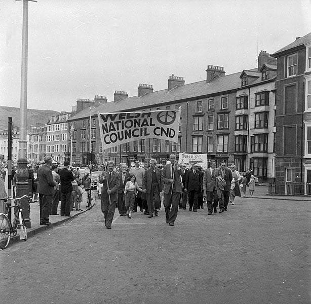 Black-and-white photo of a street march with people carrying a large Welsh National Council CND banner past tall terraced buildings