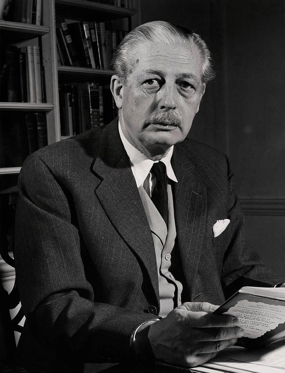 Harold Macmillan seated indoors, holding an open book with shelves of books faintly visible in the background.