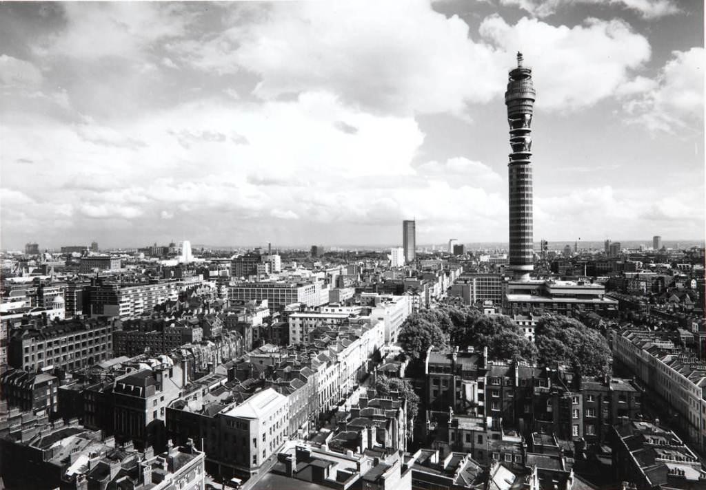 Black-and-white aerial view of central London with dense low rooftops and the tall cylindrical Post Office Tower rising prominently against a cloudy sky