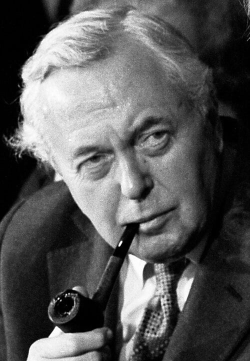 Black‑and‑white close‑up of an older man in a suit, gazing thoughtfully to the side while holding a smoking pipe to his lips in a crowded setting.