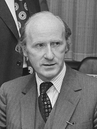 Middle-aged man in a suit and tie, seated indoors, looking slightly to the side with a serious expression, with another suited person partly visible behind.