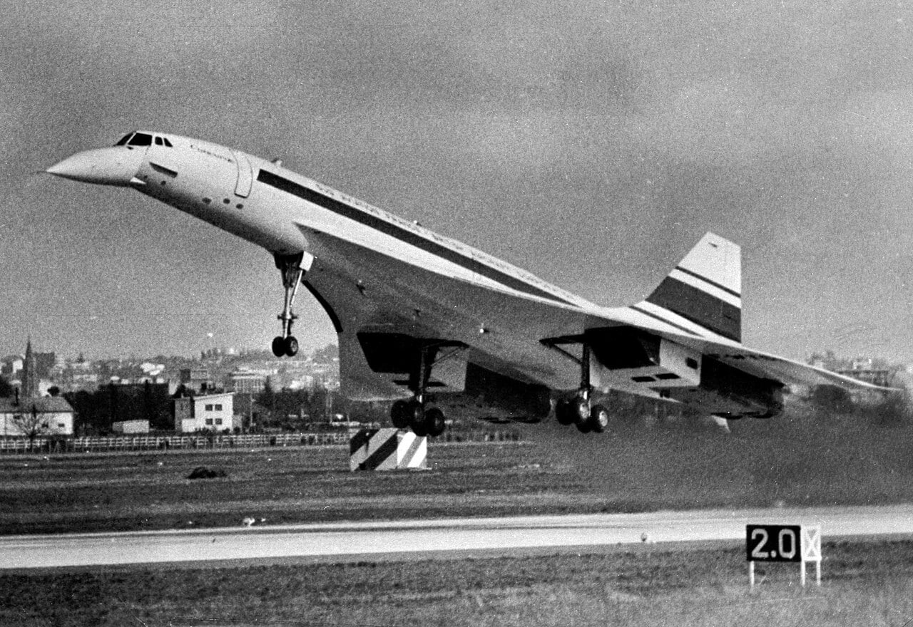 Black-and-white photo of a Concorde airliner taking off, nose raised and landing gear extended above a runway, with a cityscape blurred in the background.