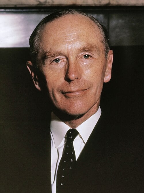 Alec Douglas-Home in a dark suit, white shirt and spotted tie, looking directly at the camera with a slight smile against a dark, softly lit background