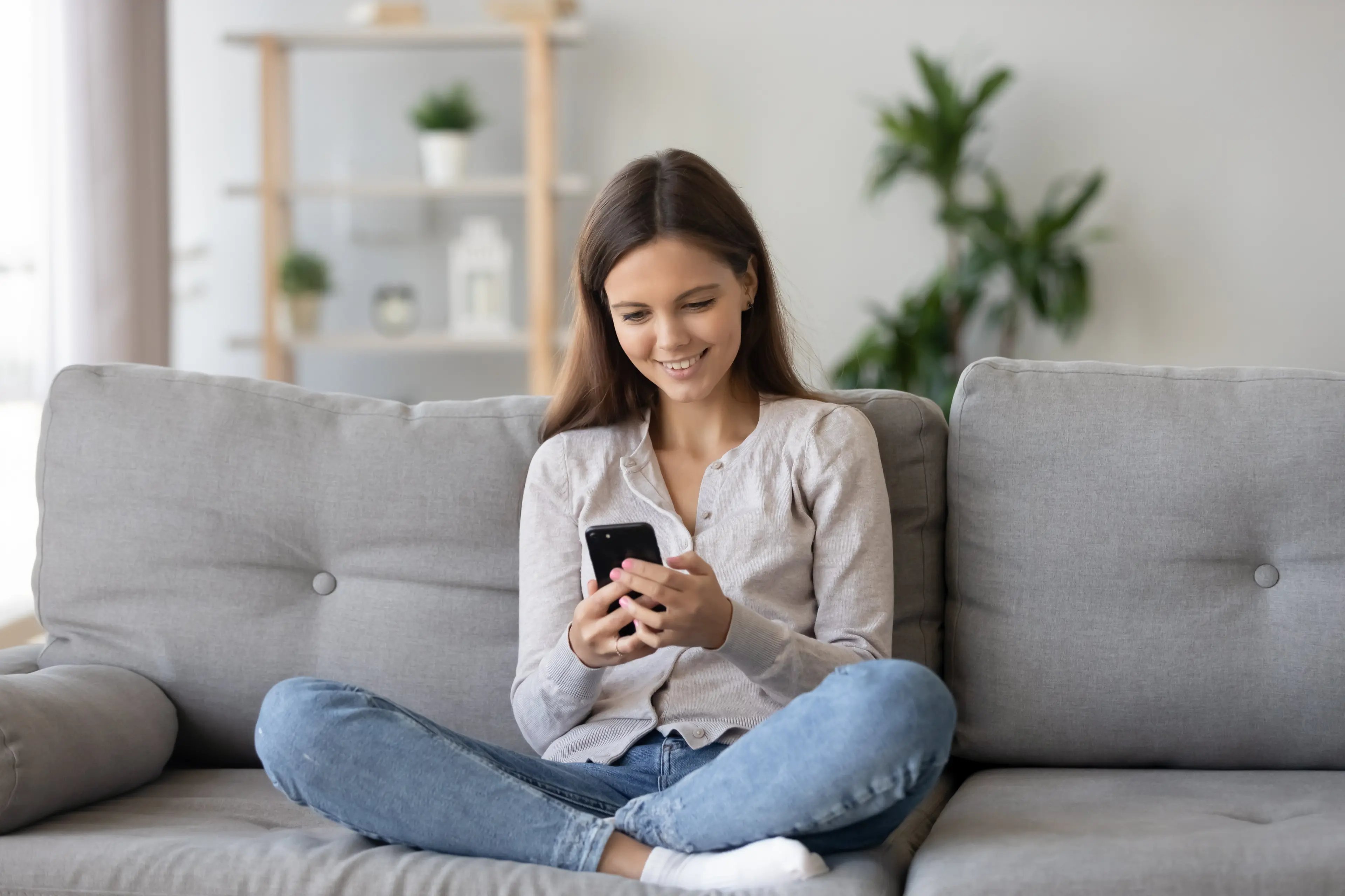 A teenage girl sitting on a sofa, smiling at her smartphone, with social media apps open on the screen.