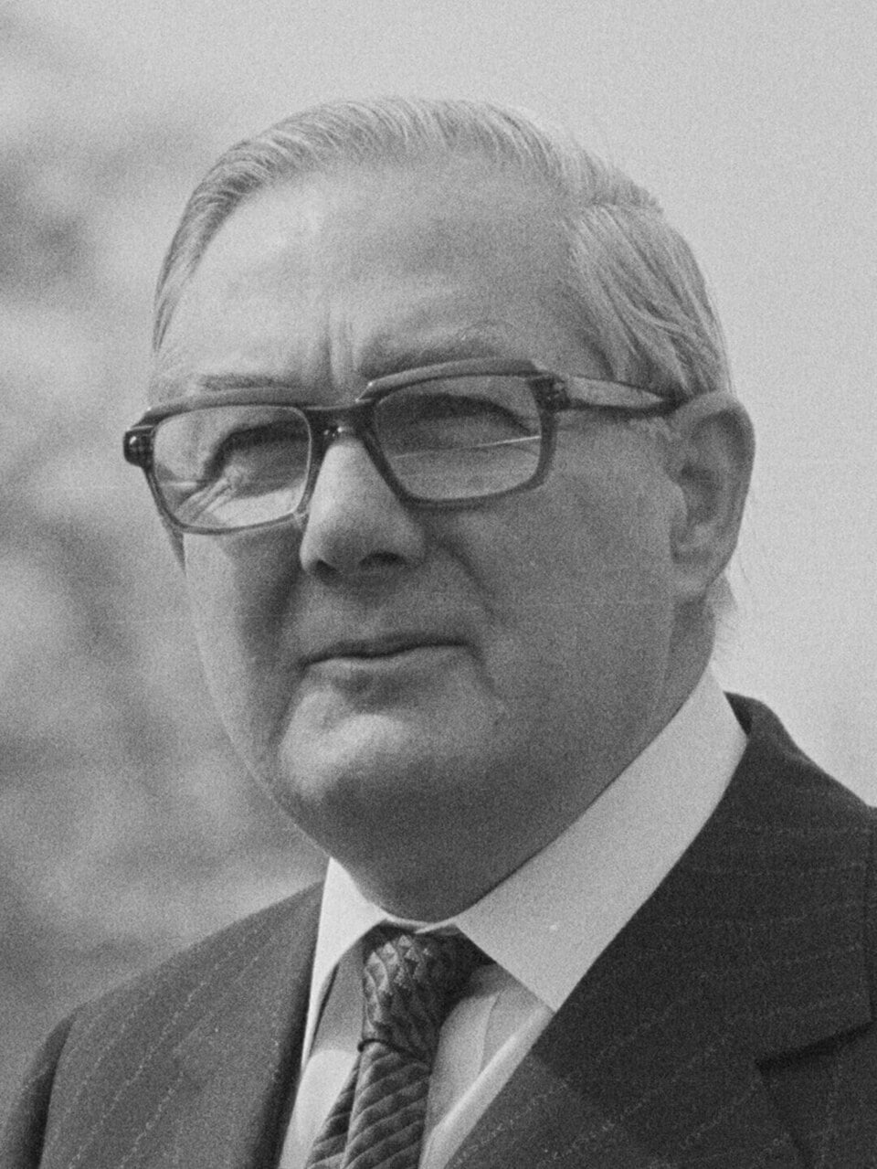 Black-and-white portrait of an older man in glasses, suit and tie, looking serious, with neatly combed hair and an out-of-focus background.