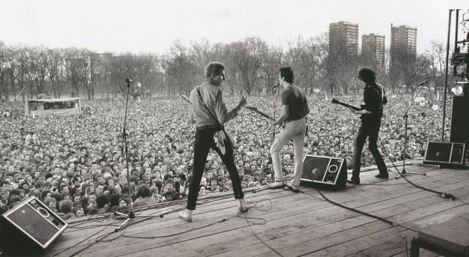 Black-and-white photo of a rock band playing guitars on an outdoor stage before a huge crowd in a park, with tower blocks and bare trees in the background