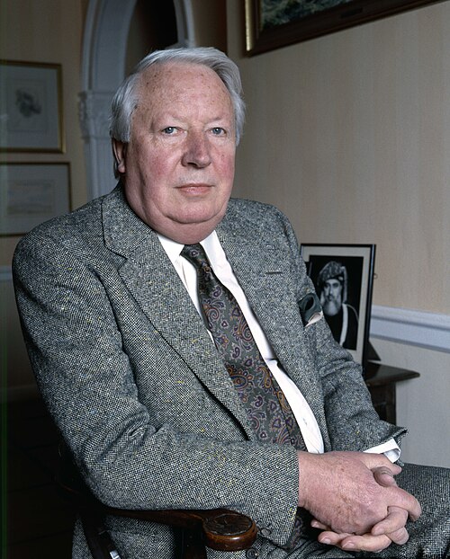 Older man in a grey tweed suit sits indoors with hands folded, looking at the camera; framed artwork and photos decorate the softly lit room behind him