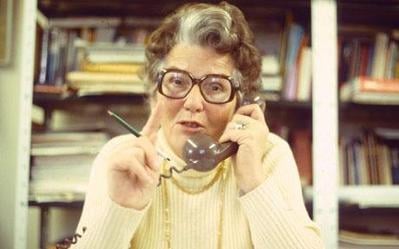 Older woman with glasses in an office, speaking on a corded telephone and gesturing with a pencil, shelves of books and papers behind her
