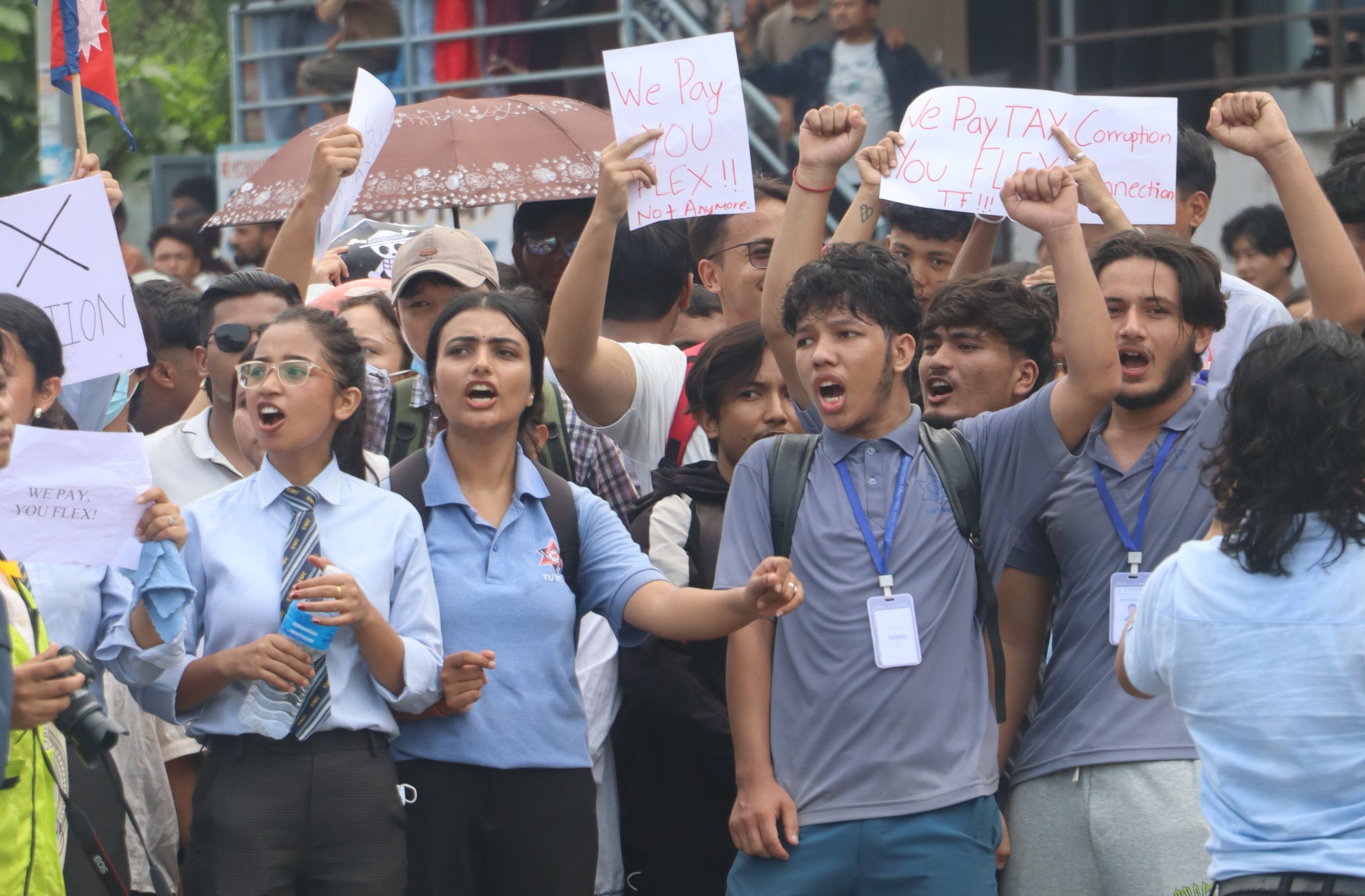 Photograph of young Nepali protesters raising fists and placards, including “We Pay You Flex!! Not Anymore” and “WE PAY YOU FLEX!”, with a Nepal flag visible at left.