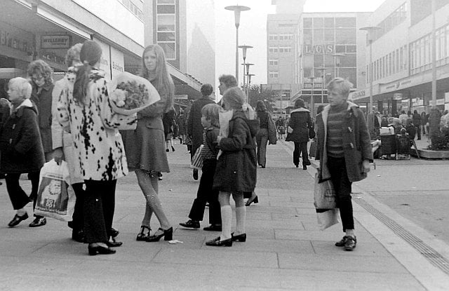Black-and-white photograph of a busy pedestrian shopping street in Harlow, with shoppers carrying bags, shopfronts, modern buildings and distinctive tall street lamps.