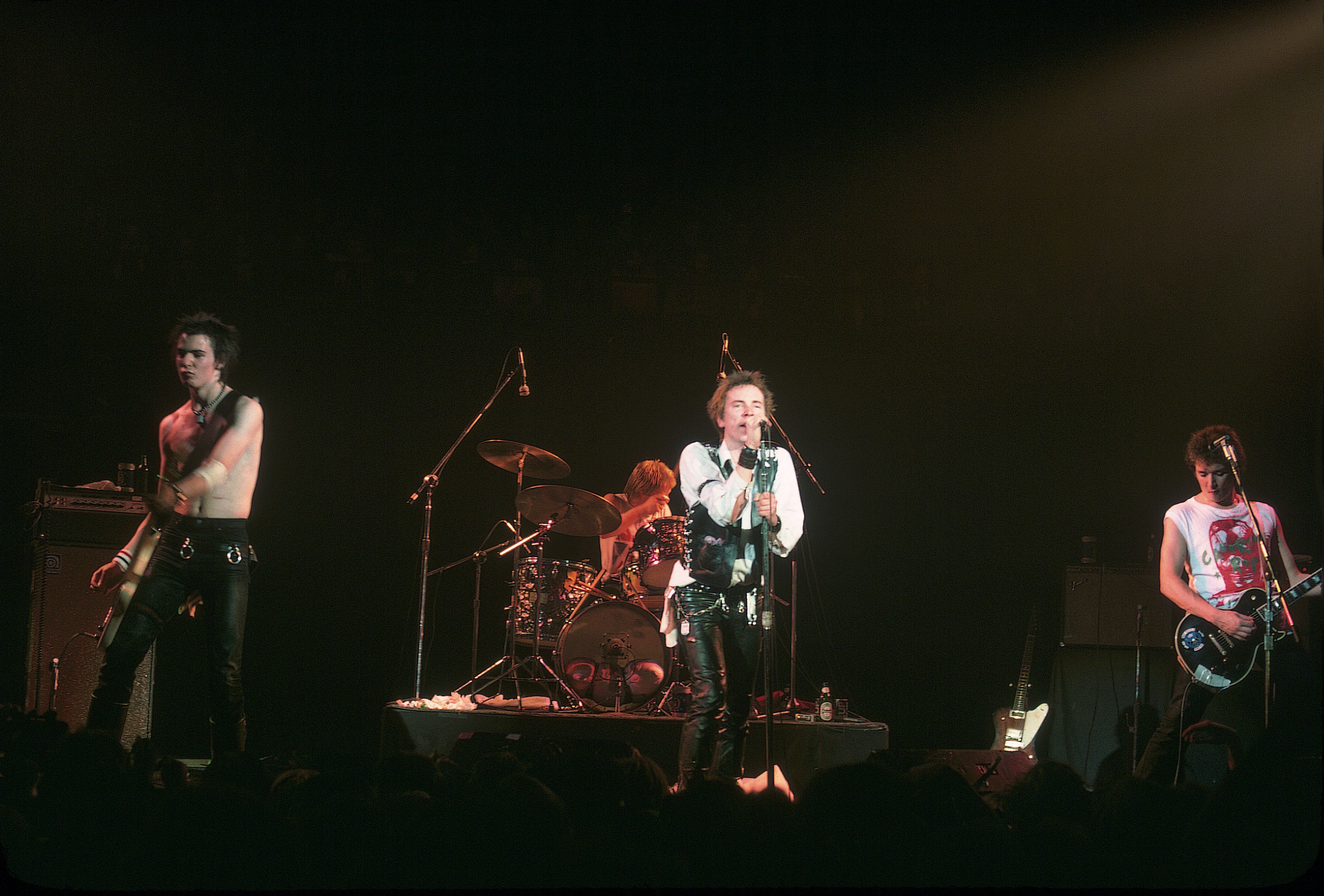 Black-and-white photo of a punk rock band performing on stage, with spiky hair, leather jackets, guitars and drums under bright concert lights