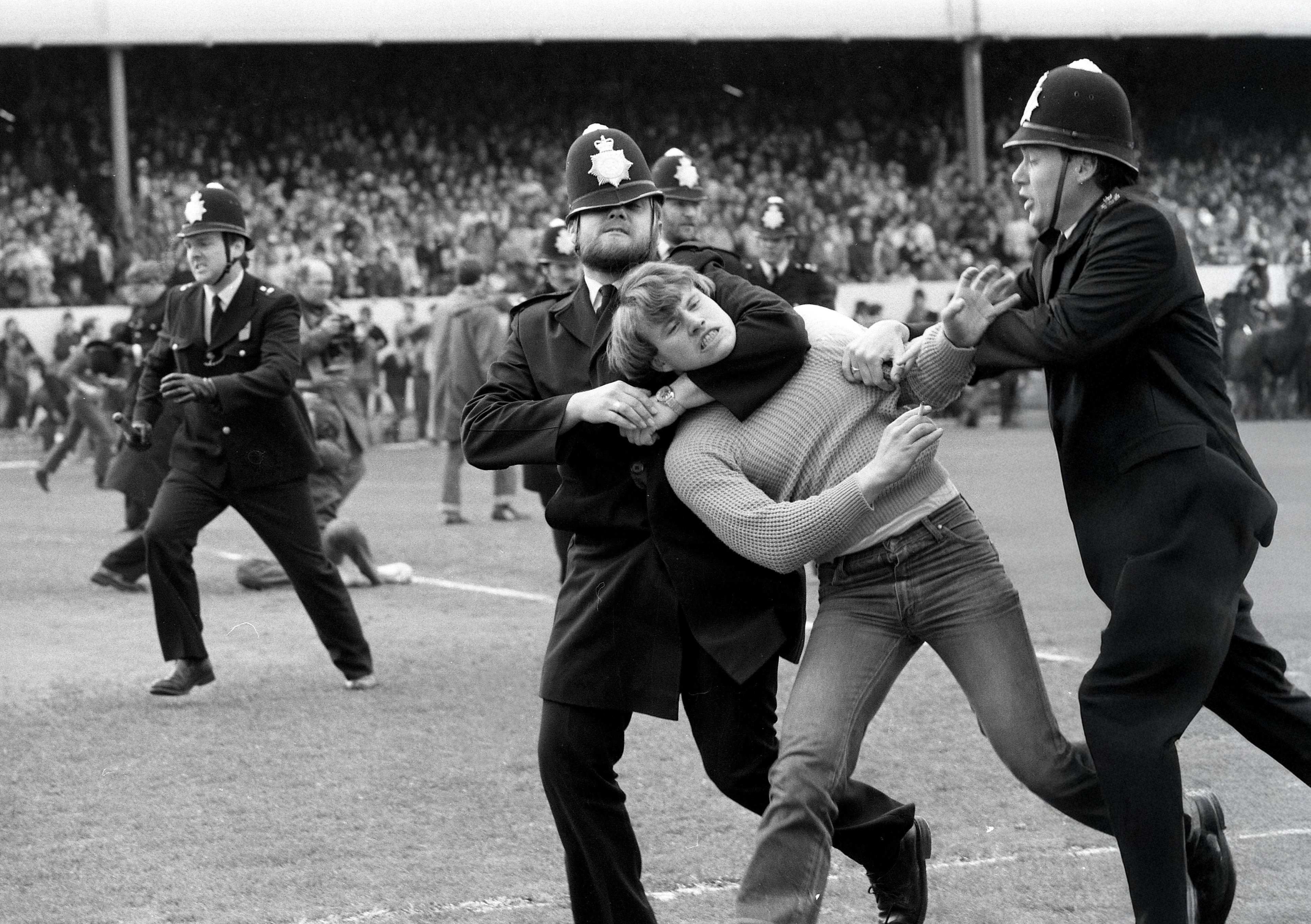 Topless football fan being led away by two policemen in uniform as a packed crowd watches from the stands in a black and white stadium scene.